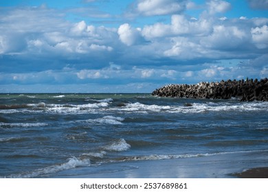 The sun shines down on calm waters as waves roll softly onto the shore, with a rocky jetty stretching into the distance beneath a sky filled with fluffy clouds. - Powered by Shutterstock