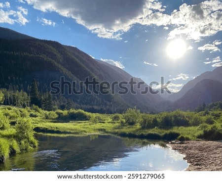  Sun shining on pine tree covered RockyMountains and wide flowing stream with lush green grasses around 