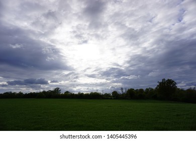 Sun shining through clouds over a green field surrounding by woodland.