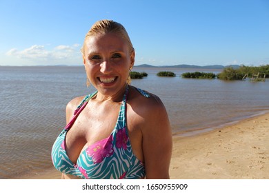 Sun tanned blond woman with blue eyes smiling at the camera with her wet hair at the beach wearing a bikini with sand water and blue sky in the background