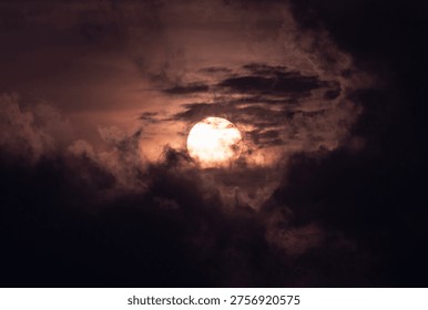 The sun is visible through dark clouds during the evening. The light from the sun creates a glowing effect on the clouds around it.