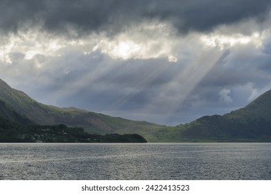 Sunbeams break through the dramatic cloud cover, illuminating parts of a serene lake surrounded by mountains as evening approaches.