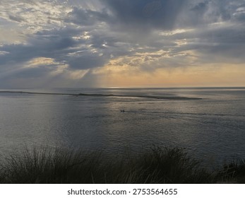 Sunbeams break through dramatic clouds over calm ocean at sunset, with distant sandbar and foreground dune grass, serene coastal seascape panorama.