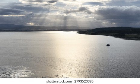 Sunbeams break through dramatic clouds over a lone boat in a tranquil Scottish bay.