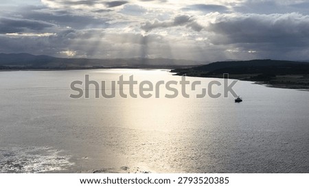 Sunbeams break through dramatic clouds over a lone boat in a tranquil Scottish bay.