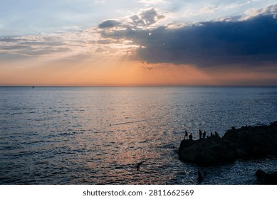 Sunbeams breaking through clouds over a calm sea with silhouettes of people on coastal rocks at sunset