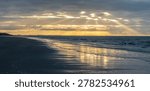 Sunbeams pierce dramatic clouds over a calm ocean reflecting golden light on the shore
