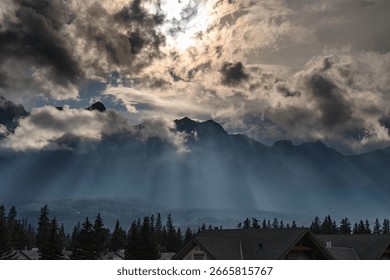 Sunbeams pierce dramatic clouds over majestic mountains and a forest, illuminating a serene landscape, sunbeams breaking through clouds over a mountainous landscape over Canadian Rockies in Canmore