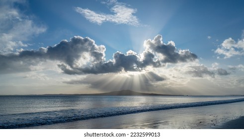 Sunbeams shining through clouds over Rangitoto Island. View from Milford beach, Auckland.