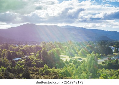 Sunbeams stream through dramatic clouds over Turangi, New Zealand, casting a magical light across the town and lush treetops with the backdrop of forested hills and distant mountain ranges