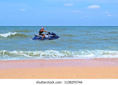 Sunburnt Man Talking By Phone Sitting On Personal Watercraft In Sea Near Empty Sandy Beach. Sunny Day, Horizon Line. 