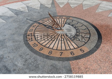 Sundial embedded into circular pattern on a tiled floor. Metal gnomon casts shadow onto dial, indicating time based on sunlight. Design includes decorative elements like crescent moon and stars 