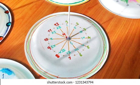 Sundial Or Solar Clock Is Is Made Of White Paper Plates With White Plastic Sticks Embroidered In The Middle, Placed On A Brown Table.