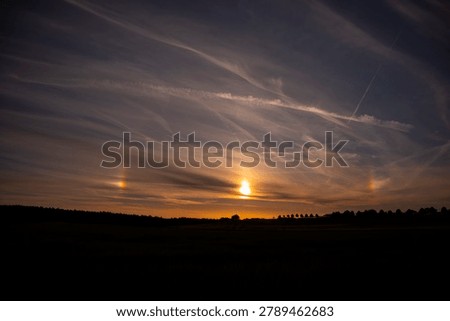 ​Beautiful sundog phenomenon during a dramatic sunset sky with wispy clouds and contrails over a dark silhouette landscape horizon.