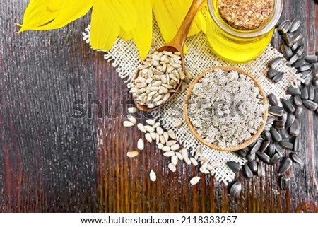 Sunflower flour in a bowl, oil in glass jar, seeds in a spoon on burlap napkin, sunflower flower on dark wooden board background from above
