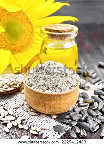 Sunflower flour in bowl, oil in glass jar, seeds in a spoon on burlap, sunflower flower on dark wooden board background