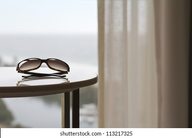 Sunglasses On A Table In A Luxury Hotel Room Or Apartment With Blurred View Of The Ocean Behind