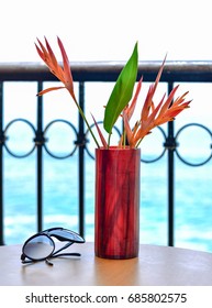 Sunglasses on a table with a red vase with bird of paradise inside with a beach and ocean background