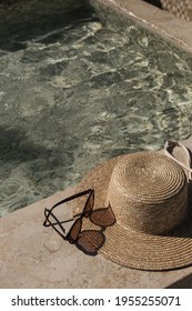 Sunglasses and straw hat on marble swimming pool side with clear blue water with waves sunlight shadow reflections. Minimal aesthetic summer vacation concept background.