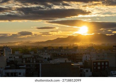 Sunlight beaming through scattered clouds illuminates the urban landscape, creating a warm, vibrant ambiance over the city buildings and distant mountain range during a golden hour sunset