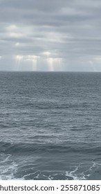 Sunlight beams cut through a cloudy sky, illuminating the calm ocean surface below.