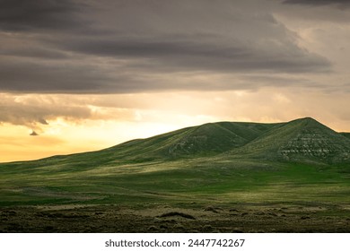 Sunlight breaks through the clouds, highlighting the sweeping landscape featuring rolling hills. The contrast between the darkened sky and the sunlit greenery creates a dramatic and serene atmosphere.