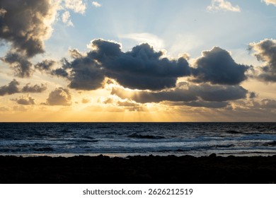 Sunlight breaks through dark clouds over a wavy ocean during sunset, casting visible rays across the sky and water with a rocky coastline in foreground.