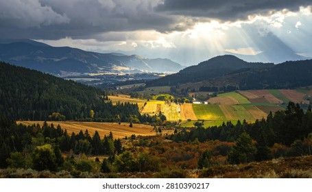 Sunlight breaks through dark clouds, illuminating a colorful valley with rolling hills and agricultural fields.