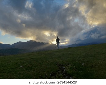 Sunlight breaks through dramatic clouds over Aitzkorri’s forested mountains near Zegama, Beunde, with a woman silhouetted in the backlight, adding mystery to the Basque landscape.