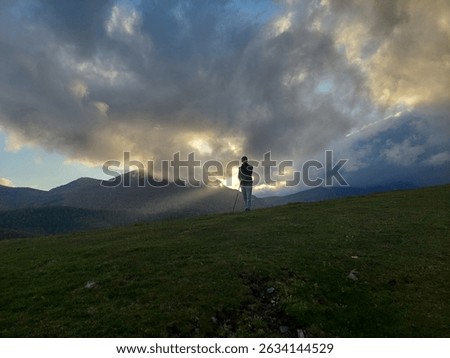 Sunlight breaks through dramatic clouds over Aitzkorri’s forested mountains near Zegama, Beunde, with a woman silhouetted in the backlight, adding mystery to the Basque landscape.