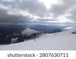 Sunlight breaks through heavy clouds over a wide mountain panorama with safety fencing and ski patrol markers near the top station at Chopok, Jasna ski resort, Low Tatras, Slovakia.