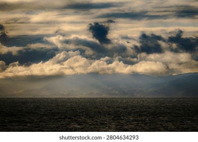 Sunlight breaks through heavy clouds casting rays over a dark, choppy sea and silhouetted mountain range