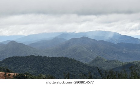 Sunlight breaks through heavy clouds, casting dramatic rays of light across layered mountain ranges that stretch toward the horizon. Dense forests blanket the rolling peaks in varying shades of green and blue, creating a striking sense of depth and scale. This awe-inspiring panoramic view captures the raw, untouched grandeur of a remote mountainous wilderness.