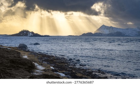 Sunlight bursts through dark clouds, casting golden rays over the tranquil waters of Gimsoy, Lofoten, Norway, creating an enchanting landscape at dusk.