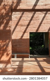 Sunlight casts dramatic diagonal shadows across the textured, layered walls of a rammed earth structure, revealing an opening to a vibrant garden