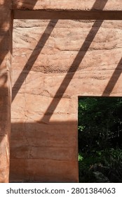 Sunlight casts dramatic diagonal shadows across a textured, layered rammed earth wall, revealing a glimpse of vibrant green vegetation beyond an architectural opening