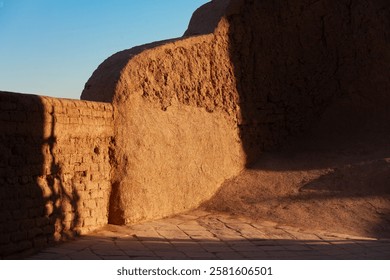 Sunlight casts dramatic shadows on the ancient mud-brick walls of a historic fortress in Iran. The warm earthy tones and textured surfaces reflect the region’s rich architectural heritage.