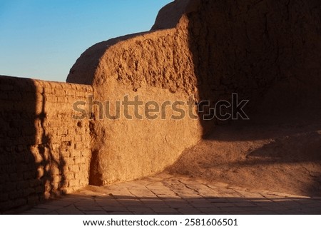 Sunlight casts dramatic shadows on the ancient mud-brick walls of a historic fortress in Iran. The warm earthy tones and textured surfaces reflect the region’s rich architectural heritage.