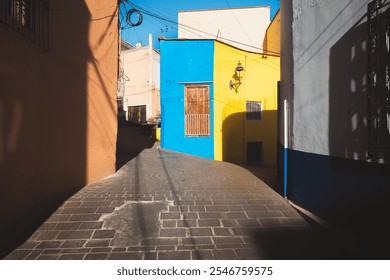 Sunlight casts dramatic shadows over colorful facades in a quaint, cobblestone alleyway in Guanajuato, Mexico, showcasing vibrant architecture and rustic charm in this historic neighborhood.