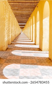 sunlight casts geometric shadows through wooden slats onto tiled corridor with yellow arches, where an unrecognisable Touareg person dressed in white traditional clothes walks at the end of the tunnel