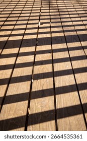 Sunlight casts a grid pattern of shadows on a wooden deck, highlighting the natural wood texture and creating an abstract outdoor background.

