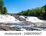 Sunlight dances on the Chutes Wilson, where the Rivière du Nord cascades vibrantly through a landscape of lush trees and weathered rocks.