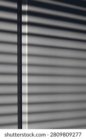 Sunlight filtering through horizontal window blinds casts sharp, geometric striped shadows onto a white wall, intersected by a dark vertical window frame. A minimalist, high-contrast abstract interior lighting composition.