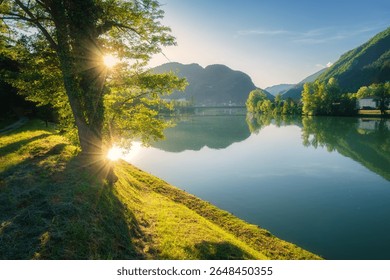Sunlight filtering through tree branches creates a beautiful scene over the calm Sava River in Slovenia, with rolling hills, gravel path, mountains and blue sky in on a serene summer sunset. Nature - Powered by Shutterstock - Get 15% off with code: PIKWIZARD15