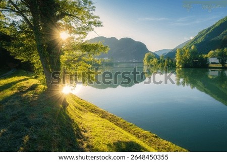 Sunlight filtering through tree branches creates a beautiful scene over the calm Sava River in Slovenia, with rolling hills, gravel path, mountains and blue sky in on a serene summer sunset. Nature