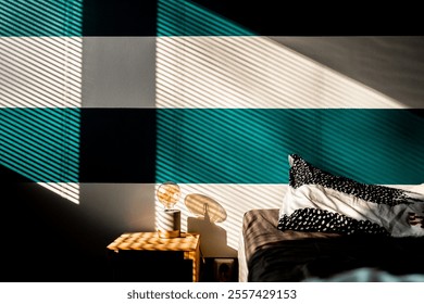 Sunlight filters through window blinds, creating striped shadows on a bedroom wall, table, and bed. A decorative pillow adds contrast, enhancing the scene's modern and peaceful aesthetic. - Powered by Shutterstock