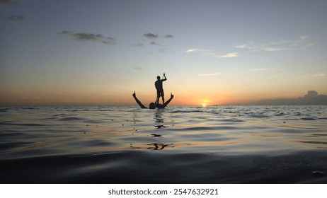 The sunlight forms a golden path on the surface of the water leading to the silhouette of a person standing on a paddle board, creating an impression of serenity against the backdrop of the sky 