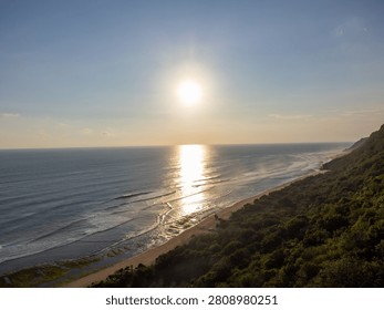 Sunlight glints off ocean waves rolling toward shore. Dunes meet dense forest along winding coastline. Silent beach holds solitary figure walking toward water. Golden hour casts long shadows over sea