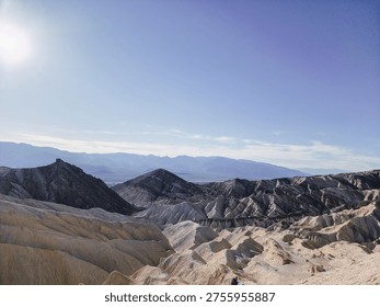 Sunlight glints over rugged, layered canyons. Desert landscape stretches beneath a clear blue sky. Distant mountains fade softly in the horizon. Vast, arid terrain invites solitude and wonder