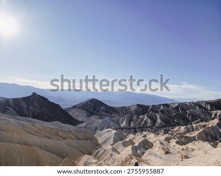Sunlight glints over rugged, layered canyons. Desert landscape stretches beneath a clear blue sky. Distant mountains fade softly in the horizon. Vast, arid terrain invites solitude and wonder